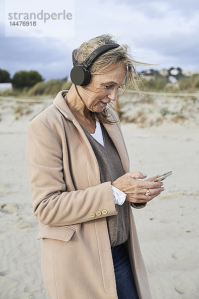 Spanien  Menorca  ältere Frau mit Smartphone und drahtlosen Kopfhörern am Strand im Winter
