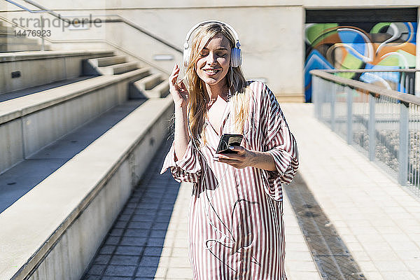 Spanien  Barcelona  Porträt einer lächelnden jungen Frau  die ein Smartphone und Kopfhörer im Freien benutzt