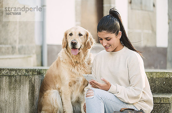 Junge Frau mit ihrem Golden-Retriever-Hund auf einer Treppe im Freien mit dem Handy