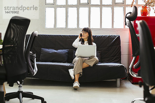 Junge Frau sitzt im Büro auf einer Couch und benutzt Handy und Laptop