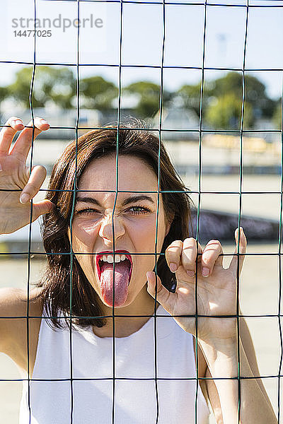 Porträt einer jungen Frau hinter einem Zaun  die ihre Zunge herausstreckt