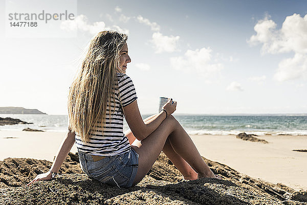 Junge Frau sitzt in einem Felsen am Strand und entspannt sich bei einer Tasse Tee