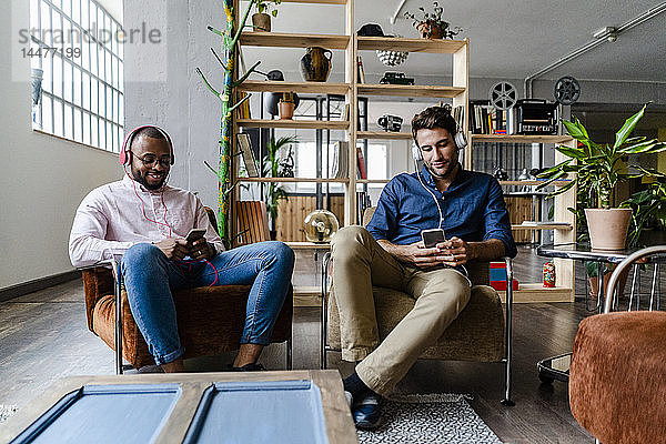 Zwei junge Männer mit Mobiltelefonen und Kopfhörern sitzen in Sesseln auf einem Dachboden