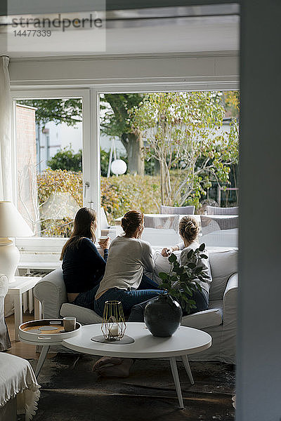 Rückansicht einer Mutter mit zwei Teenager-Mädchen  die zu Hause auf einer Couch sitzen und aus dem Fenster schauen