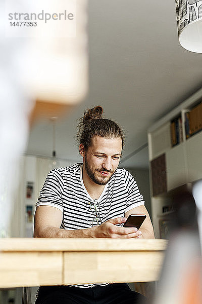 Junger Mann mit Brötchen  der zu Hause sitzt und ein Smartphone benutzt