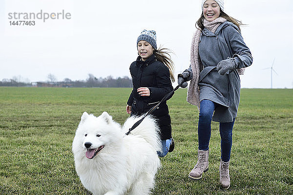 Zwei Mädchen rennen mit Hund auf einer Wiese und amüsieren sich