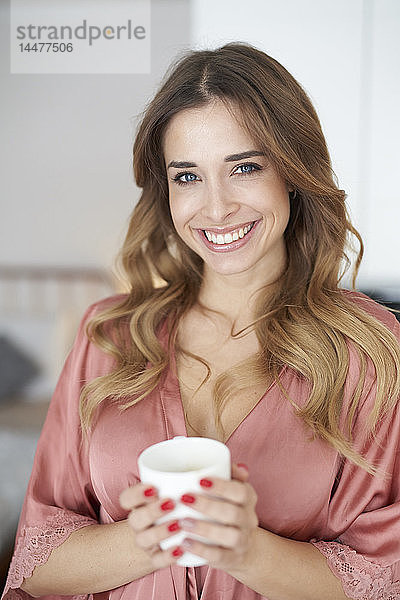 Porträt einer lächelnden jungen Frau im Bademantel mit einer Tasse Kaffee in der Hand