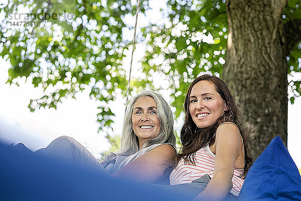 Porträt von zwei lächelnden Frauen  die sich auf einem hängenden Bett im Garten entspannen