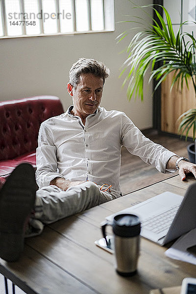 Geschäftsmann sitzt mit Füßen auf dem Schreibtisch und schaut auf den Laptop