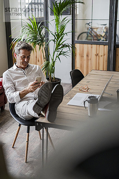 Geschäftsmann sitzt mit den Füßen auf dem Schreibtisch und benutzt ein Mobiltelefon