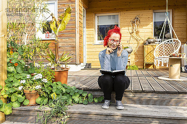 Lächelnde ältere Frau mit rot gefärbten Haaren am Telefon  die auf der Terrasse vor ihrem Haus sitzt und ein Buch liest