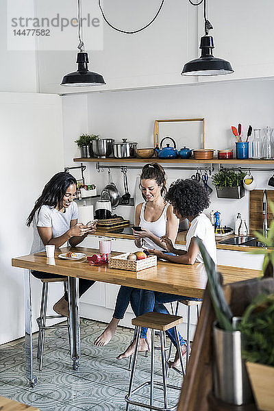Drei glückliche Frauen sitzen zu Hause am Küchentisch und benutzen Mobiltelefone