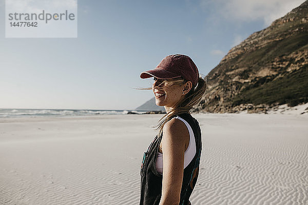 Südafrika  Western Cape  Noordhoek Beach  lächelnde junge Frau mit Basecap am Strand stehend