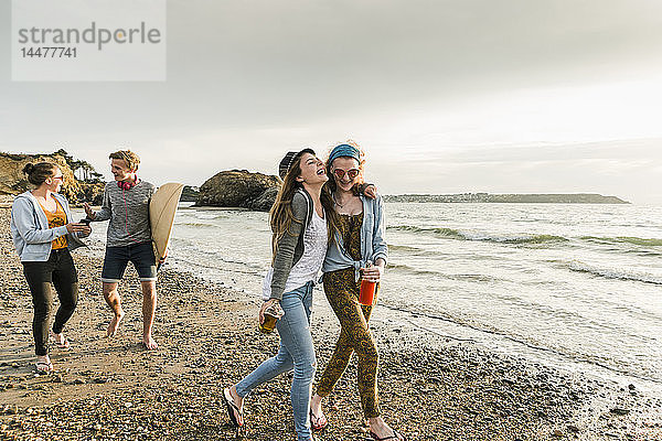 Glückliche Freunde mit Surfbrett und Getränken am steinigen Strand spazieren