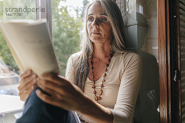 Porträt einer Frau  die zu Hause auf dem Fensterbrett sitzt und ein Buch liest