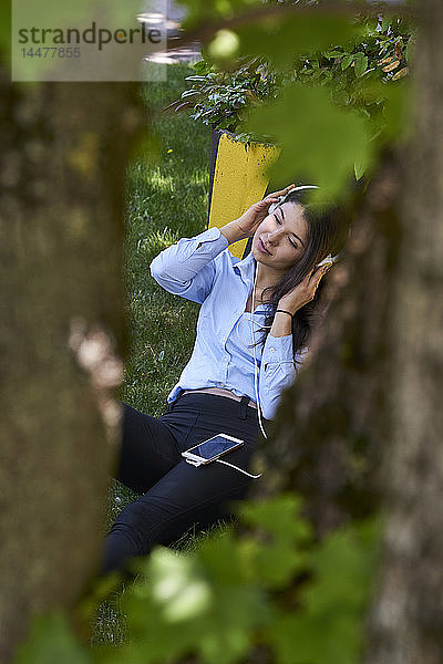 Junge Frau sitzt auf einer Wiese und hört Musik mit Kopfhörern und Handy