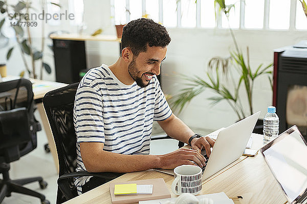 Lächelnder junger Mann mit Laptop am Schreibtisch im Büro