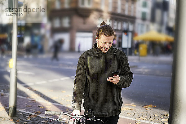 Lächelnder junger Mann mit Fahrrad in der Stadt  der auf sein Handy schaut