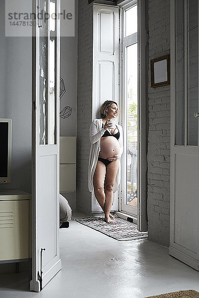 Hübsche blonde schwangere Frau steht am Fenster und hält eine Tasse Tee