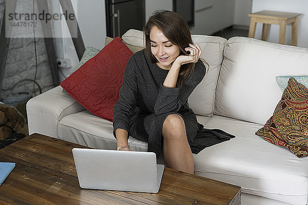 Junge Frau sitzt auf ihrer Couch und chattet auf ihrem Laptop
