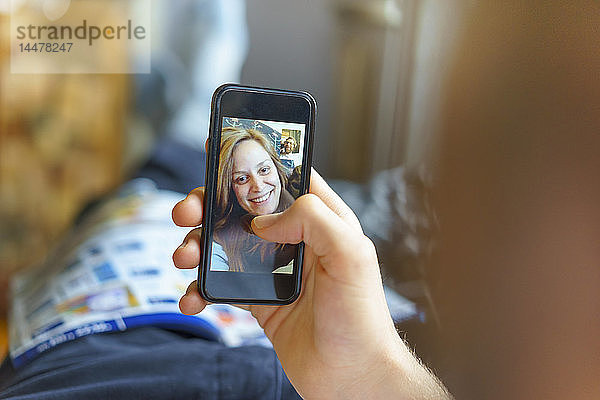 Porträt einer glücklichen jungen Frau  die mit ihrem auf der Couch liegenden Freund per Handy skypen kann