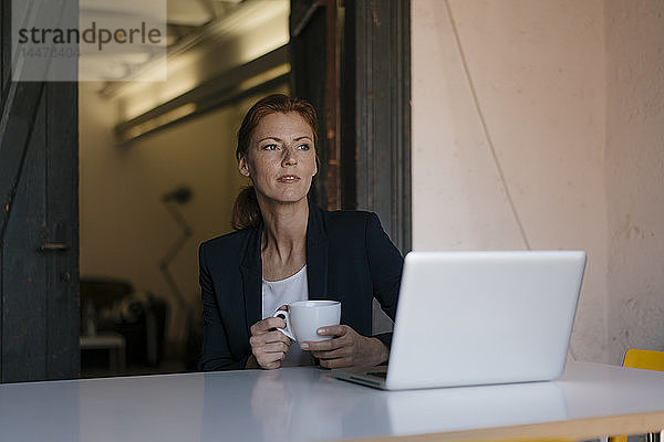 Nachdenkliche Geschäftsfrau sitzt am Schreibtisch im Büro mit Tasse Kaffee und Laptop