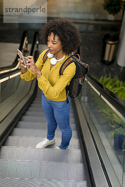 Frau mit Rucksack und Kopfhörern steht mit einem Smartphone auf der Rolltreppe