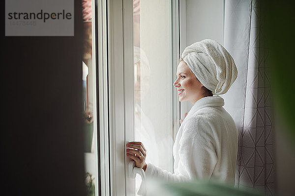 Lächelnde Frau im Bademantel mit Handtuch um den Kopf  die zu Hause aus dem Fenster schaut