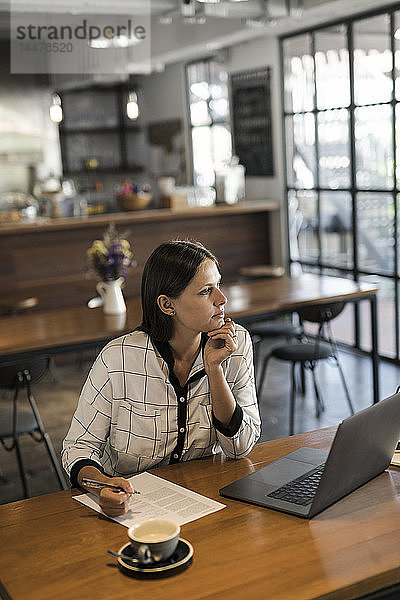 Junge Geschäftsfrau in einem Café  die auf Papier schreibt und mit einem Laptop auf einem Holztisch arbeitet