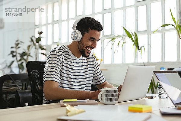 Lachender junger Mann mit Kopfhörer und Laptop am Schreibtisch im Büro