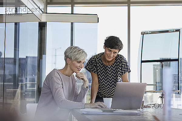 Zwei lächelnde Geschäftsfrauen teilen sich den Laptop am Schreibtisch im Büro
