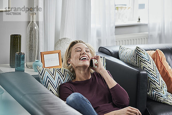 Lachende junge Frau am Handy  die zu Hause auf der Couch liegt