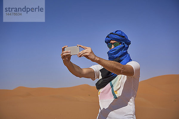 Marokko  Mann mit Sonnenbrille und blauer Tuba beim Fotografieren mit Smartphone in der Wüste