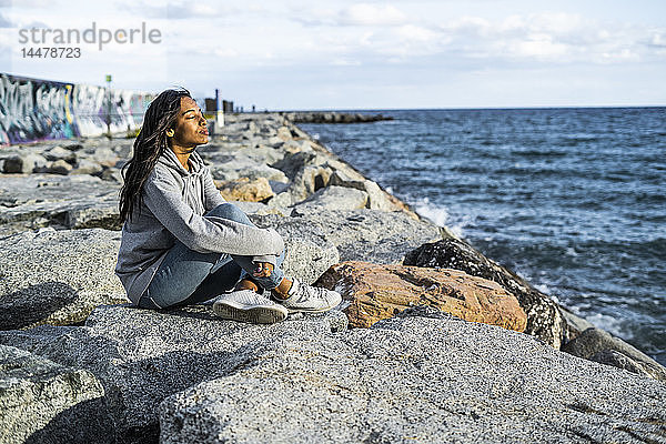 Junge Frau entspannt am Strand  auf Felsen sitzend  mit geschlossenen Augen