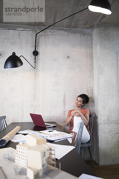 Porträt einer lächelnden Geschäftsfrau  die in einem Loft mit Dokumenten und Laptop am Tisch sitzt