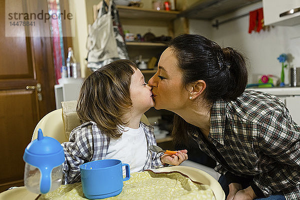 Mutter küsst Kleinkind-Sohn auf Hochstuhl in Küche