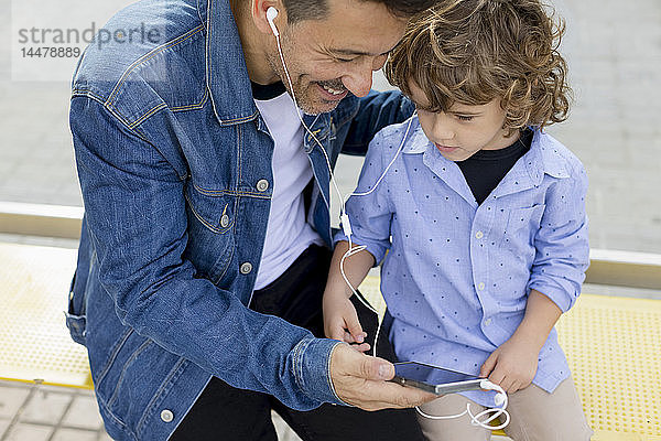 Vater und Sohn sitzen an der Straßenbahnhaltestelle in der Stadt und teilen sich Handy und Ohrhörer