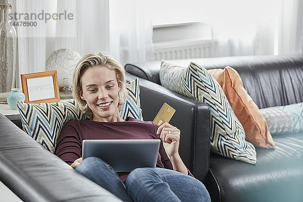 Glückliche Frau mit Tablette und Kreditkarte zu Hause auf dem Sofa liegen