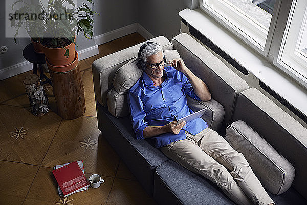 Lächelnder reifer Mann mit Tablette und Kopfhörer  der sich zu Hause auf der Couch entspannt