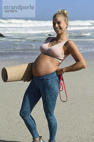 Lachende schwangere Frau hält Gymnastikmatte am Strand