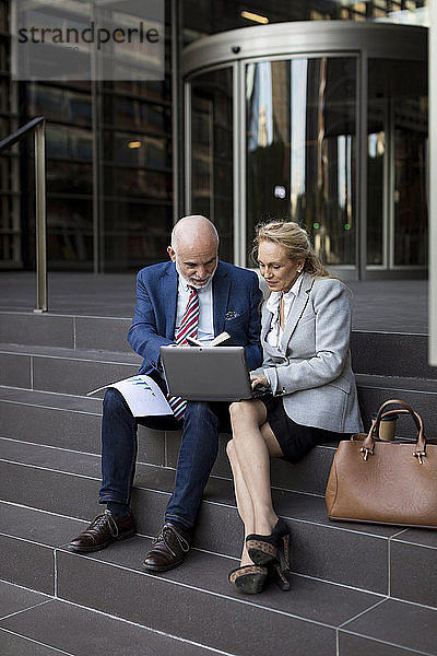 Leitender Geschäftsmann und Geschäftsfrau sitzen mit Laptop und Diagrammen auf der Treppe