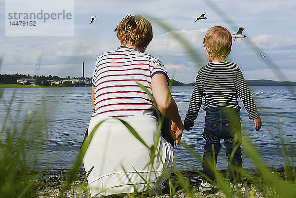 Finnland  Kuopio  Rückenansicht von Mutter und kleiner Tochter beim Beobachten von Möwen am Seeufer