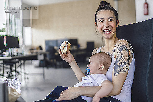 Junge Mutter mit Tätowierungen sitzt im Büro mit ihrem Baby auf dem Schoß und isst ein Sandwich