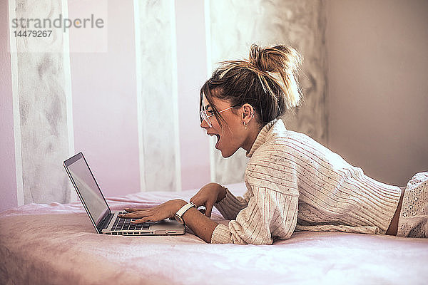 Überraschte junge Frau liegt mit Laptop auf dem Bett