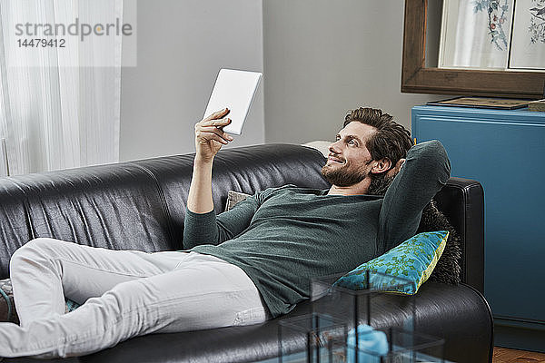 Lächelnder Mann mit Tablette  der zu Hause auf der Couch liegt