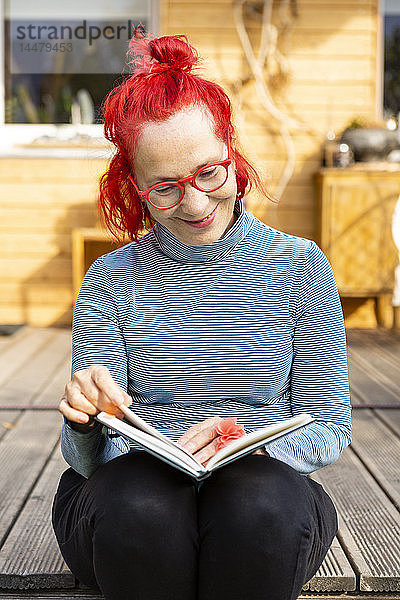 Porträt einer lächelnden älteren Frau mit rot gefärbten Haaren  die auf der Terrasse vor ihrem Haus sitzt und ein Buch liest