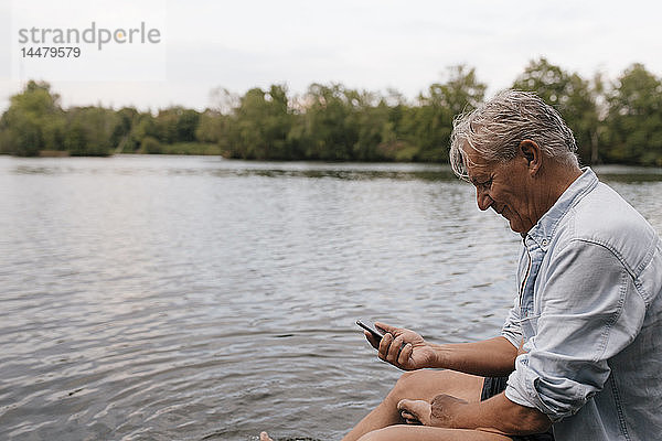 Älterer Mann sitzt mit seinem Handy an einem See