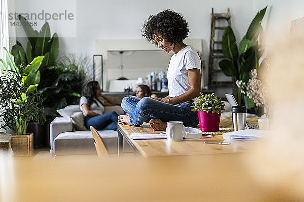 Lächelnde Frau mit Tablette auf dem Tisch zu Hause mit Freunden im Hintergrund