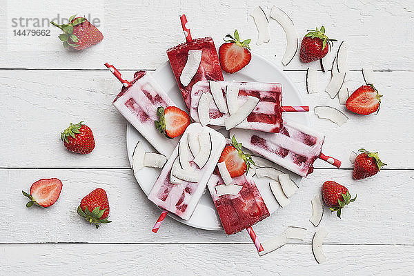 Hausgemachte Erdbeer-Kokos-Eis-Lollies mit frischen Erdbeeren und Kokosnussscheiben auf dem Teller
