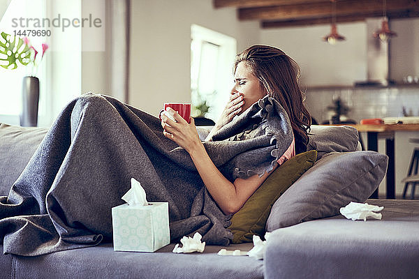 Porträt einer Frau  die krank auf dem Sofa liegt  hustet und versucht  heißen Tee zu trinken
